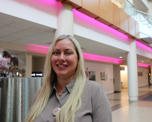 Close-up portrait of a person with long blonde hair wearing a taupe collared shirt and necklace, standing in a bright indoor atrium with wood-paneled walls, glass railings and softly blurred architectural features in the background.