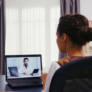 Woman participating in a telehealth video consultation with a doctor on a laptop at a home office desk.