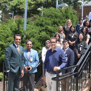 Group photo of medical residents and healthcare professionals standing on an outdoor staircase surrounded by landscaped greenery outside a healthcare facility. The team members wear business attire and identification badges while posing together on steps and walkways near the building entrance on a sunny day.
