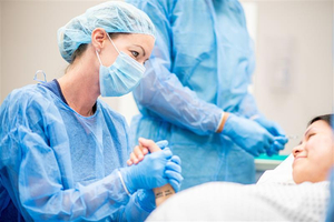 A healthcare professional wearing surgical protective equipment holds an adult patient’s hand while another clinician prepares medical supplies in a clinical procedure room.