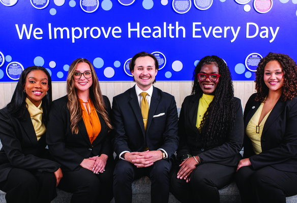 Five people in business attire seated together beneath a wall display featuring the text “We Improve Health Every Day.”