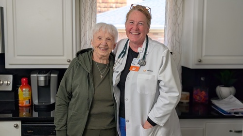 In‑home healthcare professional standing with an older adult in a home kitchen during a residential care visit.