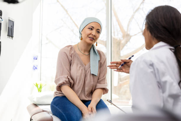 Patient wearing a head scarf sits on an exam table and speaks with a healthcare professional in a medical examination room with large windows.