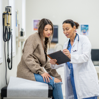 Healthcare professional reviewing information on a tablet with a patient seated on an examination table in a medical clinic.