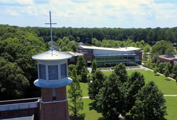Aerial view of a brick tower topped with a white cross overlooking a landscaped campus with red-brick buildings, glass-fronted structures, walking paths, green lawns, and surrounding trees under a partly cloudy sky.
