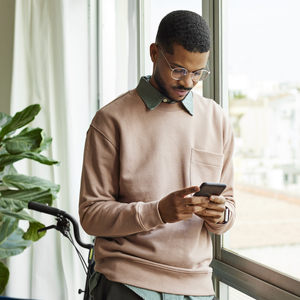 An adult stands indoors beside large windows, looking down at a smartphone while a bicycle rests nearby in a bright, modern interior space.