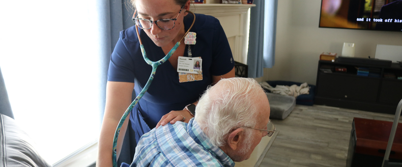 A nurse uses a stethoscope to listen to an older patient's back while the patient sits in a chair at home.