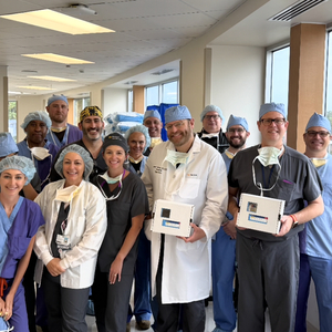 A group of 12 healthcare professionals poses together in a brightly lit hospital hallway lined with large windows. The team wears surgical scrubs in shades of blue, gray, and purple, along with surgical caps and some masks hanging around their necks. Two members near the front hold small white medical supply boxes, while another wears a white physician's coat.