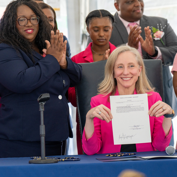 Virginia Governor Abigail Spanberger signs the Queen Candis Sickle Cell Act of Virginia during a ceremonial bill signing at the Sentara Community Care Center in Newport News, Thursday, June 11, 2026. Family members of Candis Gabriella King, elected officials, healthcare leaders, and community advocates stand behind her applauding, with Virginia and U.S. flags and a tribute display honoring Candis visible in the background.