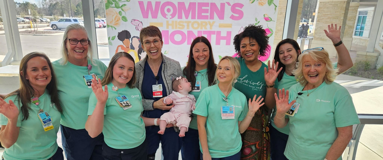 Nine labor and delivery healthcare workers pose in front of a Women's History Month banner at Sentara Albemarle Medical Center. The middle worker is holding an infant.