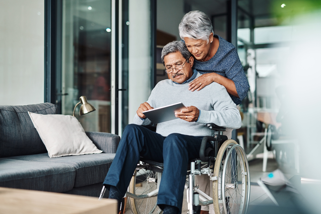 Two adults in a modern living room, one seated in a wheelchair using a tablet while another stands close behind with hands on the seated person’s shoulders, both looking at the screen. A gray sofa, floor lamp, and glass doors are visible in the background.