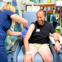 A healthcare worker wearing gloves administers an injection to a seated patient in a hospital treatment room. Medical equipment and monitors are visible in the background.