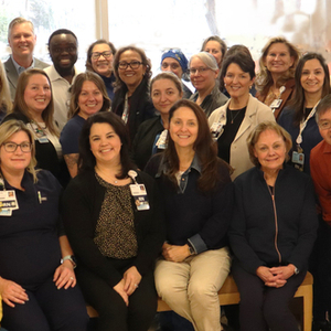 Members of Sentara Northern Virginia Medical Center’s MRSA prevention team gather for a group photo inside the hospital. Healthcare professionals and support staff wearing scrubs, business attire, and identification badges stand and sit together in a lobby area near large windows. A Magnet with Distinction sign is visible in the background.
