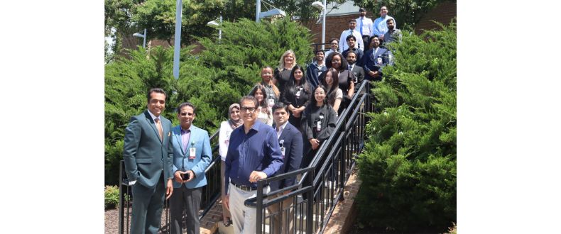 Group photo of medical residents and healthcare professionals standing on an outdoor staircase surrounded by landscaped greenery outside a healthcare facility. The team members wear business attire and identification badges while posing together on steps and walkways near the building entrance on a sunny day.