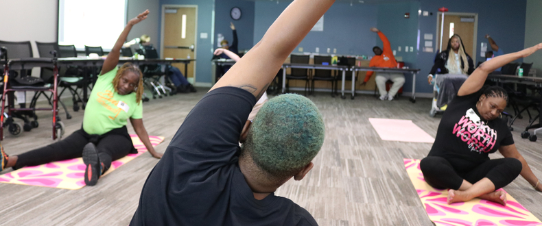 Participants in a support group session sit on yoga mats in a conference room while practicing seated stretching exercises, with one participant in the foreground leading a side stretch.