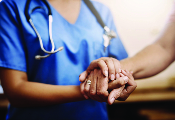 A healthcare worker wearing scrubs gently holds another person's hand in a supportive manner, shown in a close-up view.