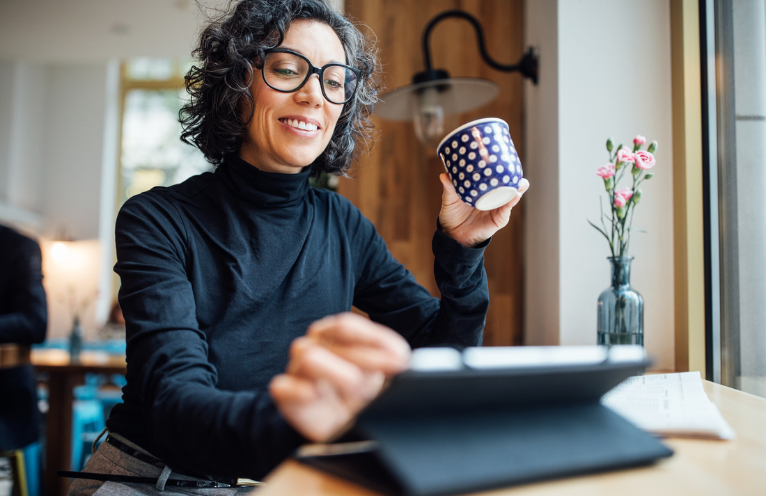 Person with short curly hair wearing glasses and a black turtleneck sits at a table, holding a blue patterned mug while using a tablet. A glass vase with pink flowers and a lamp are visible in the softly lit background.