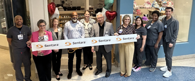 Sentara pharmacy staff and hospital leaders gather for a group photo holding a ceremonial Sentara-branded ribbon in front of the new Sentara To Home outpatient pharmacy at Sentara Northern Virginia Medical Center.