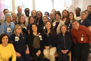 Members of Sentara Northern Virginia Medical Center’s MRSA prevention team gather for a group photo inside the hospital. Healthcare professionals and support staff wearing scrubs, business attire, and identification badges stand and sit together in a lobby area near large windows. A Magnet with Distinction sign is visible in the background.