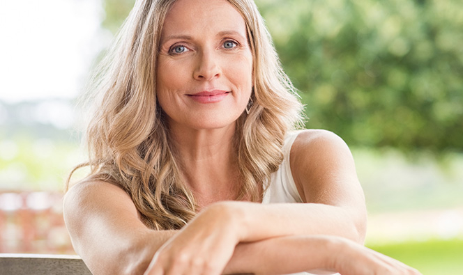 An adult sits outdoors with arms resting on a bench, posing calmly in a green, sunlit setting.
