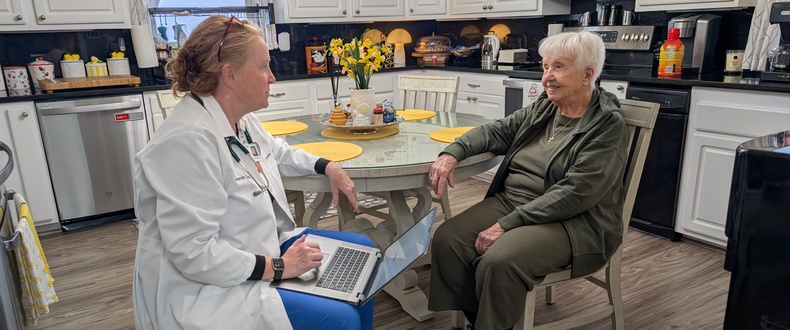 Healthcare professional seated with an older adult at a kitchen table, using a laptop during an in‑home visit.