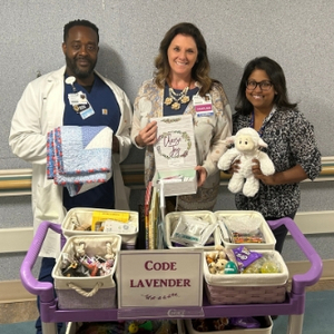 Three healthcare team members stand behind a purple Code Lavender support cart in a hospital hallway. The cart contains comfort items, books, sensory tools, snacks, a stuffed lamb, and a folded quilt used to support patients, families, and caregivers during stressful situations. A sign on the cart reads “Code Lavender.”