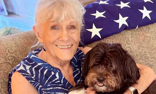 An older woman with light blonde hair pulled back and bangs smiles while sitting on a patterned beige couch, holding a small, fluffy brown dog with white paws and a lighter-colored muzzle. She wears a sleeveless blue-and-white patterned dress, and a folded American flag with white stars is draped over the back of the couch behind her.
