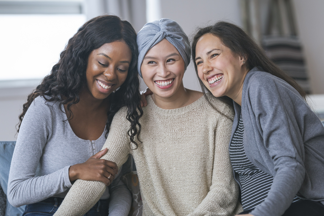 Three people sit close together on a couch indoors, leaning toward one another. The person in the center wears a light-colored sweater and a head wrap, while the people on either side wear casual tops and sweaters. All three face toward the camera in a softly lit living room setting.