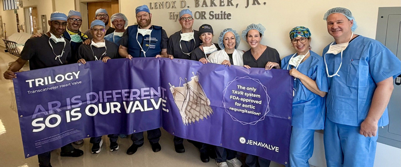 The Sentara Structural Heart Team poses in a hospital hallway in surgical scrubs and caps, holding a large purple JenaValve banner marking the first commercial use of the Trilogy® Transcatheter Heart Valve system — the only FDA-approved TAVR for aortic regurgitation — in Virginia. The sign behind them reads "Lenox D. Baker, Jr. Hybrid OR Suite."